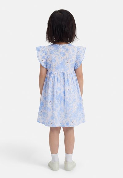 Παιδί που φοράει ανοιχτό μπλε φόρεμα με λουλουδάτο σχέδιο και βολάν στα μανίκια, λευκές κάλτσες και λευκά παπούτσια, στέκεται γυρισμένο με την πλάτη προς τα εμπρός σε λευκό φόντο.