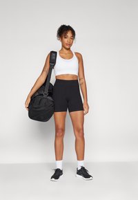 Sort atletisk shorts, hvid crop top, holder en sort sportstaske. Letvægtsmateriale, tætsiddende design med teksturerede detaljer på tasken.