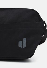 Bolsa de tela negra con una textura suave, que presenta un sutil detalle del logotipo azul. El diseño es rectangular y cuenta con un cierre de cremallera.