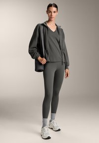 Tummanharmaa urheiluvaatetus, johon kuuluu vetoketjuhuppari, tiukat legginsit ja valkoiset urheilukengät. Mukana musta laukku, jossa on joogamatto.