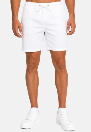 Modèle masculin portant un short blanc uni avec cordon et des baskets blanches, debout devant un fond neutre.