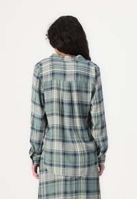 Long-sleeved plaid shirt in muted green, featuring white and navy lines, a back yoke detail, and button cuffs with a rounded hem.