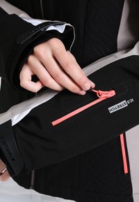 Main tirant la fermeture éclair rose sur la manche noire et grise d'une veste avec les étiquettes visibles ICEPEAK et ICESTRETCH.