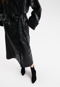 Cappotto trench in pelle nera con design doppiopetto, vita regolabile con cintura e maniche lunghe. Abbinato a stivali neri alla caviglia.
