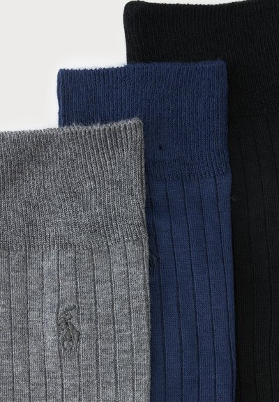 Trois chaussettes côtelées pliées en gris, bleu marine et noir, avec un petit logo brodé sur la chaussette grise.