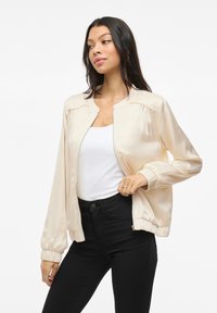Ljust beige satin bomberjacka med ribbade manschetter, dragkedja och slät textur, kombinerad med en vit topp och svarta jeans.