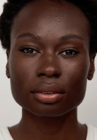 Primer plano de un rostro femenino de piel oscura, con piel suave, pómulos definidos y cabello corto y texturizado. Maquillaje natural con labios suaves.