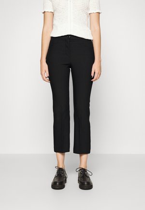 Femme portant un pantalon noir droit court, des chaussures noires lacées et un haut blanc à manches courtes texturé, debout devant un fond uni.