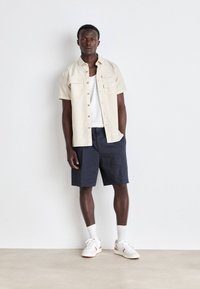 Camicia beige con bottoni e due tasche sul petto, canotta bianca, pantaloni corti blu navy e sneakers bianche con accenti arancioni, in piedi su uno sfondo neutro.