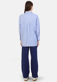 Camicia oversize a righe blu e bianche con yoke posteriore, maniche lunghe, abbinata a pantaloni larghi blu navy e calzature bianche.