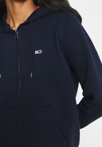 Felpa con zip di colore navy e cappuccio con corde, con un piccolo logo ricamato sul petto e una tasca frontale. Texture morbida, design casual.