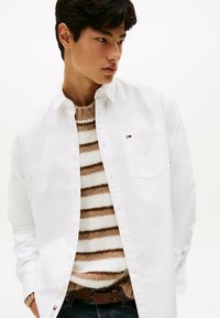 Young man wearing an unbuttoned white shirt over a brown and white striped knit sweater, dark jeans, and a brown belt against a plain background.