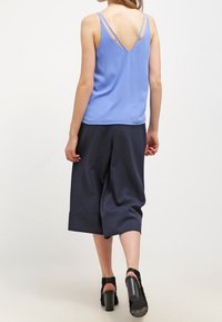 Světle modrý saténový top na ramínka s tenkými pásky, kombinovaný s tmavými kalhotami culottes a černými sandály na blokovém podpatku, pohled zezadu.