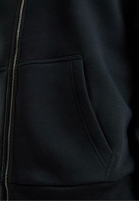Sweatshirt à zip noir en tissu doux. Il dispose d'une poche avant et de poignets côtelés. Texture lisse avec un design simple et épuré.