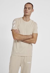 Beige t-shirt med korte ærmer lavet af blødt stof, med tre hvide diagonale striber på venstre skulder og rund halsudskæring.