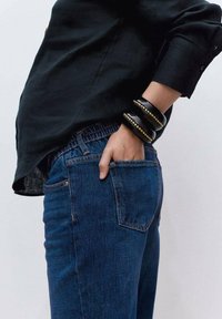 Denim jeans in dark blue with a fitted design, featuring visible stitching and a front pocket. Worn with a black shirt and stacked bracelets.