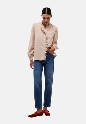 Chemise beige à manches longues avec plastron et poignets à volants, associée à un jean bleu à jambe droite et des ballerines rouges. Tenue simple et décontractée.