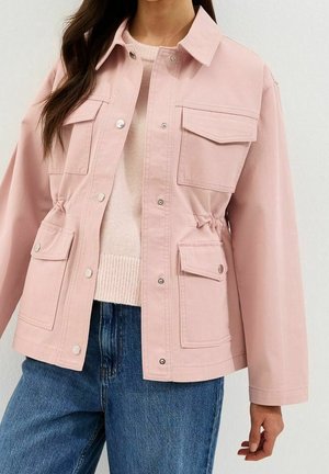 Femme portant une veste rose clair à boutons avec plusieurs poches à rabat sur un pull beige et un jean bleu.