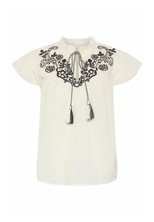 Blouse blanche à manches courtes avec broderie florale noire sur la poitrine et lien au col avec des pompons noirs et blancs.