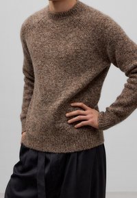 Brązowy sweter o fakturowanym splocie, z okrągłym dekoltem i prążkowanymi mankietami. Noszony na ciemnych plisowanych spodniach, prezentuje luźny krój.