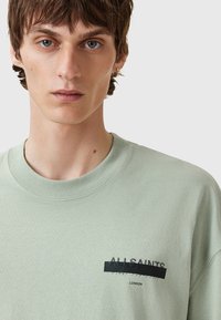 Camiseta de algodón verde claro con cuello redondo, mangas cortas y un parche negro con el logo "All Saints" en el lado izquierdo del pecho.