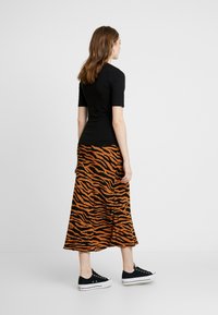 Schwarzes Kurzarmoberteil kombiniert mit einem langen, orange- und schwarzgestreiften Rock im Tiger-Muster und schwarzen Turnschuhen mit weißen Sohlen.