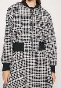 Veste bomber à carreaux noir et blanc avec une fermeture éclair à l'avant, des poignets côtelés et une taille ajustée. Tissu texturé avec un motif dessiné.