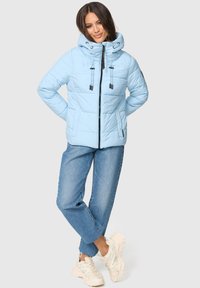 Lichtblauwe pufferjack met een hoge kraag, verstelbare trekkoordkap en rits aan de voorkant; gecombineerd met blauwe jeans en lichte sneakers.