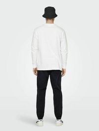 Wit T-shirt met lange mouwen en een rechte snit, gecombineerd met donkere broek en een zwarte bucket hat. Model staat met de rug naar de camera en laat een casual look zien.