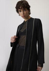 Crna jakna dugih rukava sa srebrnim detaljima na zatvaraču; složena preko prozirne crne bluze. Tkanina ima gladak tekstur, sa minimalističkim dizajnom.