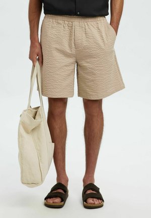 Beige, gestructureerde shorts met een elastische tailleband, gecombineerd met een canvas draagtasje en donkere sandaal. De shorts hebben zijzakken.