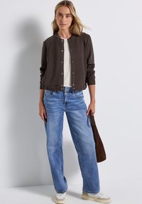 Brun kort bomberjacka med tryckknappar, bärs över en vit topp, ihop med ljusblå vida jeans och en brun axelväska.