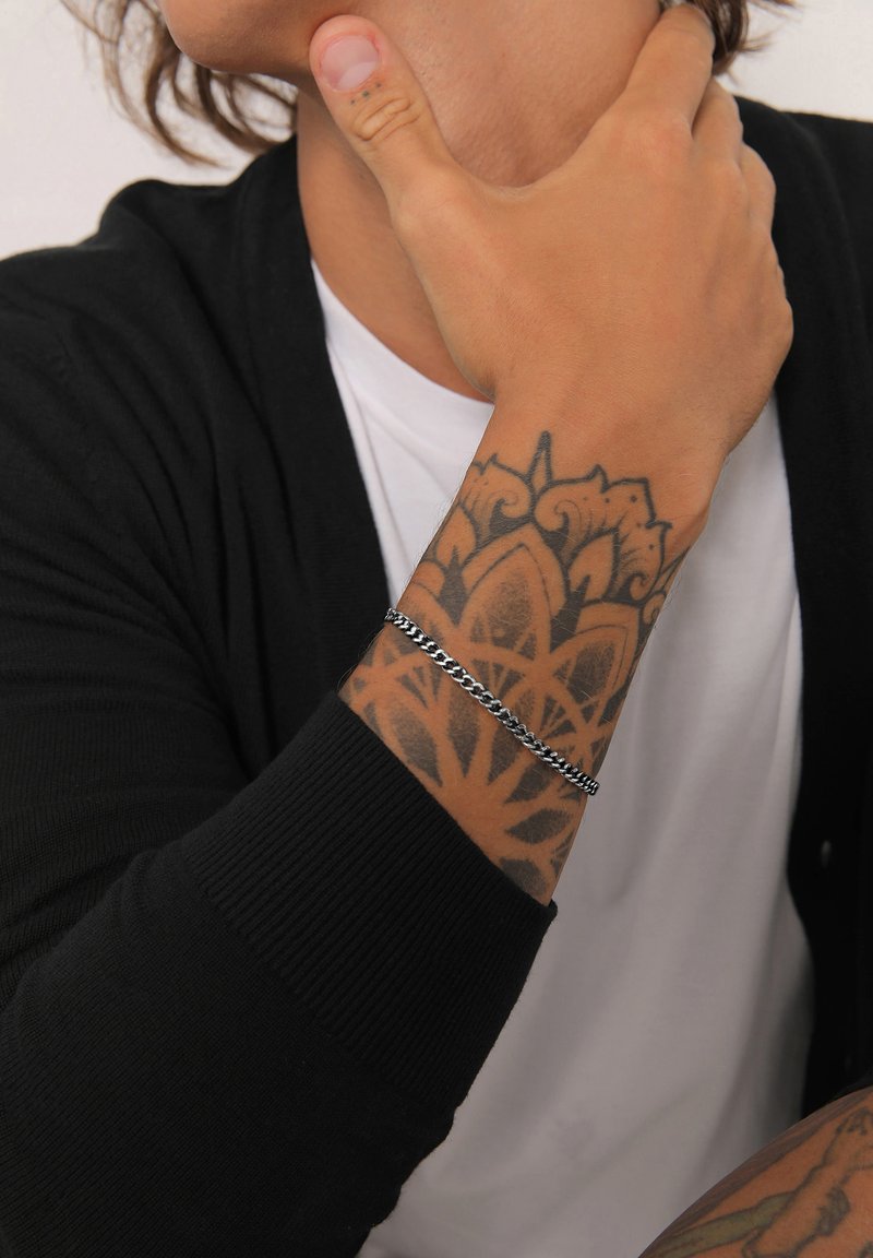 Silver chain bracelet on a tanned wrist with a detailed black tattoo. Worn with a black cardigan over a white shirt.