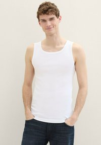 Débardeur blanc à côtes avec un col rond, de coupe ajustée. La modèle l'associe avec un jean en denim foncé, se tenant avec les mains dans les poches.