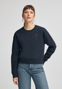 Marinblå kort sweatshirt i bomull, med ribbad halsringning och muddar, samt en liten röd logotyp på bröstet. Kombinerad med ljusblå jeans.