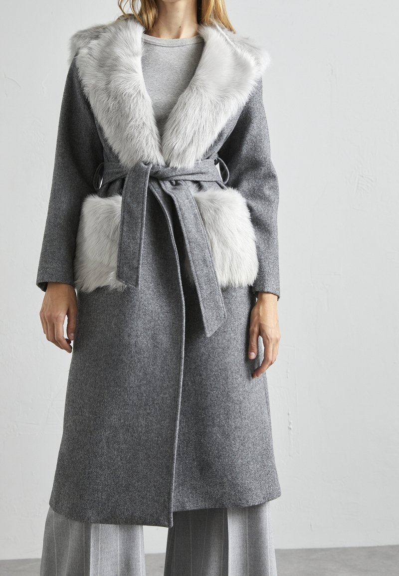 Woman wearing a long grey coat with a wide white faux fur collar and matching fur pockets, tied at the waist with a belt.