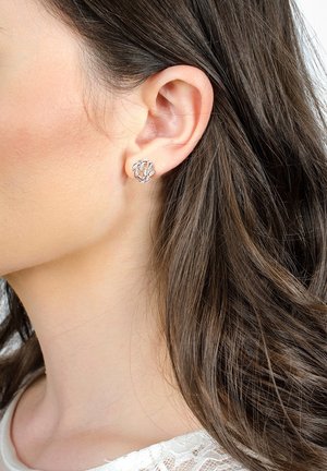 Oreille gauche d'une femme avec une petite boucle d'oreille argentée en forme de feuille délicate, cheveux brun foncé, et un haut en dentelle blanche visible.
