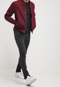 Bómber burdeos, camiseta negra, vaqueros negros y zapatillas blancas. La chaqueta tiene un escote redondo y un cierre con cremallera.