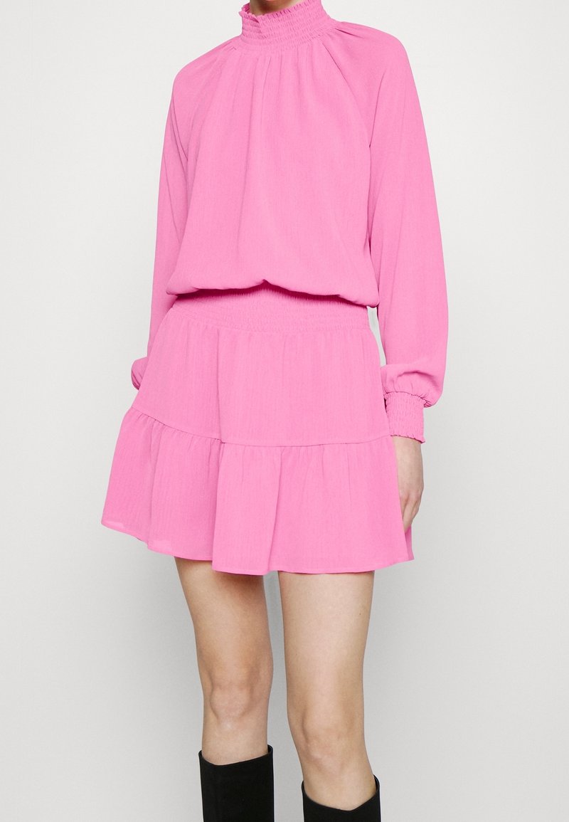Femme portant une robe longue rose vif à manches longues avec un col et une taille froncés, associée à des bottes noires montant jusqu'au genou.