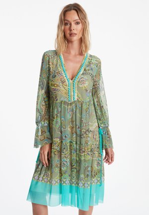 Grünes, gemustertes Kleid mit V-Ausschnitt, langen Ärmeln und rüschgesäumtem Saum. Verfügt über dekorative türkisfarbene Akzente und einen halbtransparenten Overlay.