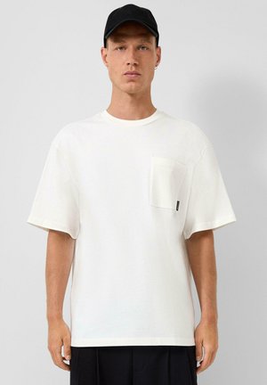 Uomo con maglietta bianca a maniche corte con taschino sul petto e cappellino nero, in piedi davanti a uno sfondo grigio chiaro uniforme.