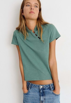 Young woman wearing a short-sleeve green top with button details and blue denim jeans, standing with hands in pockets against a white background.