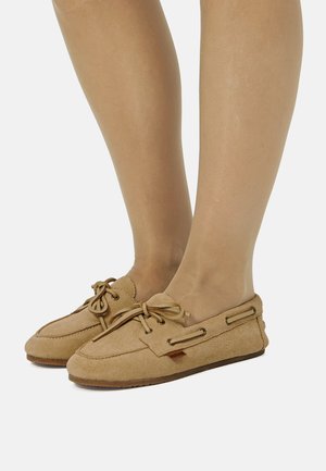 Pieds portant des chaussons mocassins en daim beige avec des lacets noués en nœuds, présentés sur un fond clair uni.