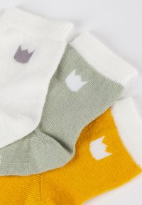 Drei Paar Socken: Weiß mit violettem Krönchen, Grün mit weißem Krönchen und Gelb mit weißem Krönchen, alle mit einem gerippten Bündchen.
