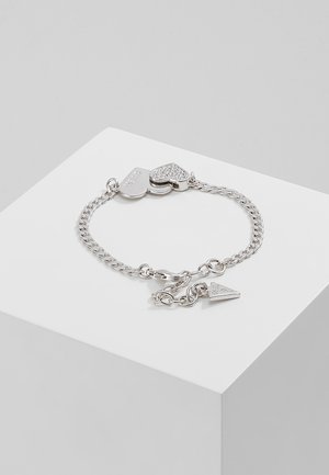 Silbernes Kettenarmband mit zwei herzförmigen Anhängern, einer mit kleinen Kristallen besetzt, dargestellt auf einem weißen Podest vor grauem Hintergrund.