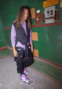 Jeune personne avec des dreadlocks portant une chemise violette, un gilet noir, une veste nouée, un pantalon noir et des baskets colorées, debout contre un mur métallique vert.