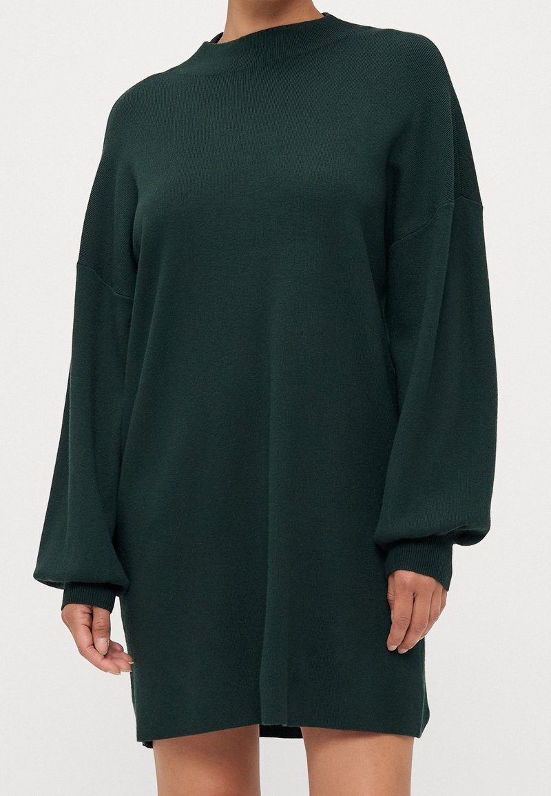 Robe pull en tricot vert foncé à manches longues portée par une personne avec des poignets détendus et un col rond.