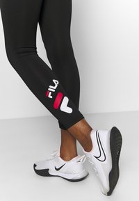 Schwarze Leggings mit einem weißen und rosa "FILA"-Logo am Unterschenkel. Getragen mit weißen Nike-Schuhen, die ein schwarzes Swoosh-Design aufweisen.