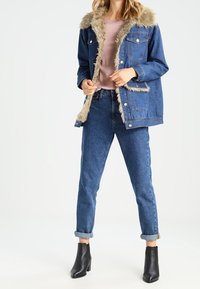 Žena v modré džínové bundě s béžovým kožešinovým lemováním, růžovém topu, modrých ohrnutých džínách a černých kotníkových botách, stojící před bílým pozadím.