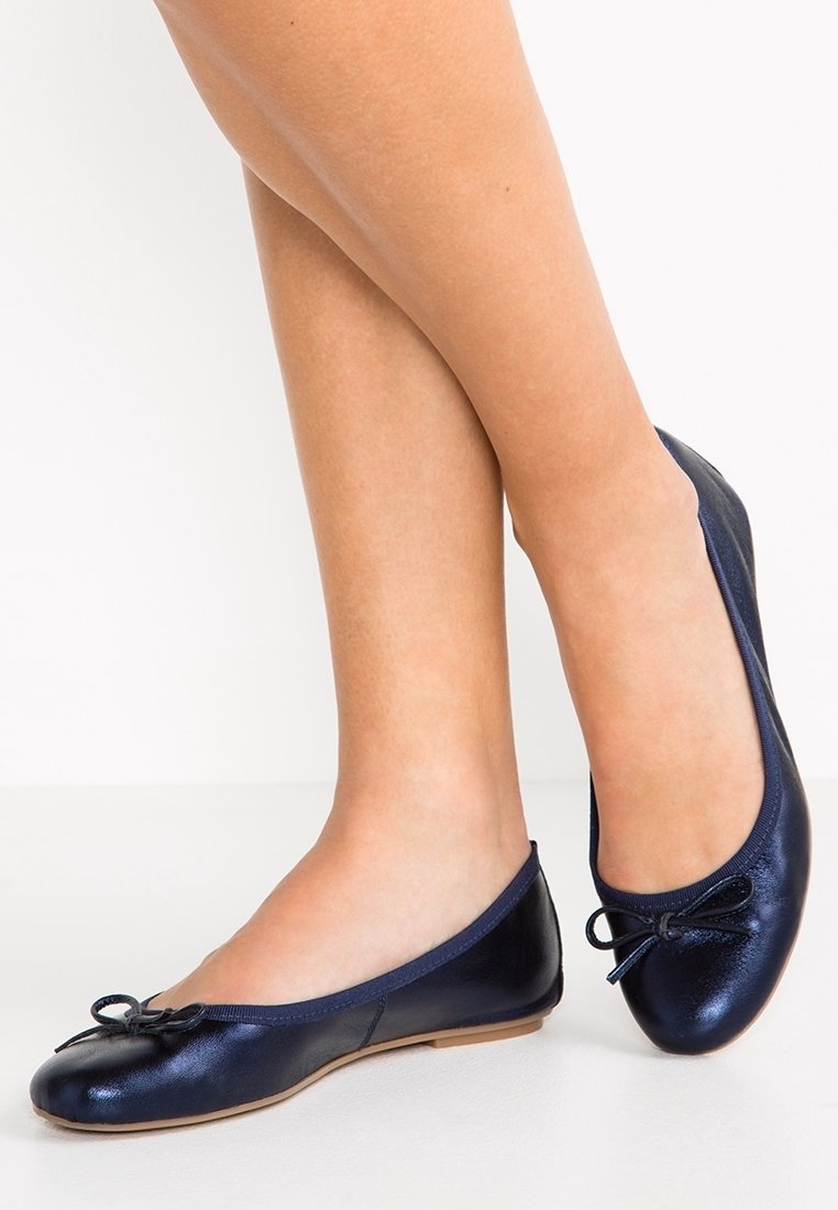 Jambes portant des ballerines bleu marine brillantes avec de petits nœuds, debout sur une surface blanche unie.
